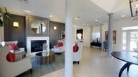 This single-story space at Stewards Crossing showcases a thoughtfully designed open floor plan with a refined contemporary aesthetic. The living area serves as a focal point, anchored by a sleek fireplace set against a charcoal gray accent wall with elegant panel molding detail. A large circular gold-framed mirror above the fireplace adds a decorative touch, complemented by a wall sconce that enhances the sophisticated atmosphere. Structural columns define the space while maintaining an open, airy feel throughout. Light neutral flooring flows seamlessly across the main living area, contributing to a bright and cohesive interior. Recessed lighting is installed throughout the ceiling, providing consistent, modern illumination. Tasteful furnishings, including wingback chairs with bold red accent pillows and a latticed round coffee table, highlight the stylish design sensibility. In the background, a glimpse of what appears to be a kitchen or bar area with pendant lighting adds functional depth to the layout. Large windows dressed with sheer curtains allow natural light to filter in, enhancing the overall warmth and openness of the space. The combination of neutral tones, architectural details, and curated furnishings creates a polished and inviting environment.