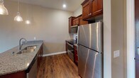 This single-floor residence at 2117 Abbey Rd in Onalaska, Wisconsin offers a well-appointed, open-concept layout designed for comfortable everyday living. The kitchen serves as a central focal point, featuring rich dark wood cabinetry, granite countertops, a double-basin stainless steel sink set into a peninsula island, and a full suite of stainless steel appliances including a refrigerator, range, and built-in microwave. A trio of pendant lights hang above the peninsula, providing both task lighting and a stylish design detail. The kitchen flows seamlessly into the living and dining area, which is finished with warm hardwood-style flooring throughout. A sliding glass door at the far end of the living space leads to a private balcony, allowing natural light to fill the room. The bedroom is carpeted for comfort, features a ceiling fan with light fixture, and receives natural light through a window with blinds. The full bathroom includes tile flooring, a vanity with dark wood cabinetry and stone-look countertop, a large mirror, and a full soaking tub with shower combination. The overall ambiance is clean, modern, and neutral throughout.