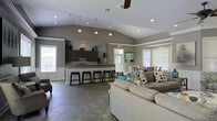 Located at 2100 Farm Springs Rd in Summerville, South Carolina, this single-story community clubhouse offers an open-concept layout designed for comfortable gathering and socializing. The space is anchored by soaring vaulted ceilings that lend an airy, expansive feel throughout the main area. Recessed lighting and a ceiling fan work in tandem to keep the environment well-lit and comfortable.

The full kitchen is a standout feature, equipped with dark cabinetry, granite-style countertops, stainless steel appliances, and a generous bar counter lined with sleek barstools — ideal for casual dining or entertaining. Pendant lights hang above the kitchen area, adding warm accent lighting.

The open living area flows seamlessly from the kitchen, furnished with a large sectional sofa and coordinating accent pieces in neutral tones accented by pops of teal and gold. Multiple windows fitted with blinds allow abundant natural light to fill the space while maintaining privacy. A dining area is visible toward the rear, providing additional seating for residents and guests.

Durable large-format tile flooring runs throughout, offering a clean, low-maintenance aesthetic that ties the entire space together. The overall ambiance is welcoming, modern, and functional — well-suited for community events and everyday resident use.