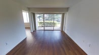 This single-floor space is defined by a clean, minimalist aesthetic that prioritizes natural light and an open feel. The main living area is anchored by warm, rich hardwood flooring that runs throughout, lending a sense of continuity and warmth to the space. A large sliding glass door dominates the primary wall, offering an expansive view of mature trees and well-maintained shrubbery outside. This generous glazing floods the interior with abundant natural light, creating a bright and welcoming atmosphere throughout the day. Sheer curtain panels frame the sliding door, allowing for privacy without sacrificing the connection to the outdoors. The walls are finished in a crisp white, complementing the natural tones of the flooring and enhancing the sense of openness. A secondary window on the adjacent wall further contributes to the well-lit environment. The layout flows openly between areas, with a subtle transition visible toward an adjoining room, suggesting a practical and straightforward floor plan. The overall ambiance is calm and understated, offering a neutral canvas well-suited for a variety of interior styles and furnishing arrangements.