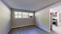 This efficiently designed 2-bedroom, 1-bathroom single-floor unit offers a well-organized layout that maximizes every square foot. The space is finished with neutral-toned walls and clean white trim throughout, creating a cohesive and tidy aesthetic.

The primary bedroom is the largest of the two, featuring wide-plank hardwood-style flooring, a textured ceiling, a wall-mounted air conditioning unit, and a baseboard heating system. A bank of windows along one wall lets in generous natural light and offers views of surrounding greenery. The second bedroom is carpeted and includes a sliding-door closet, a baseboard heater, and its own window with outdoor views.

The kitchen stands out as a clear highlight of the unit, showcasing rich dark-wood cabinetry, granite-style countertops, stainless steel appliances including a dishwasher and range, and a window above the sink that brings in natural light. Light hardwood-style floors complete the kitchen's polished look.

The bathroom is cleanly finished with floor-to-ceiling white tile surrounding a tub-shower combo, a marble-style tile floor, and a wood vanity topped with a granite countertop and a framed mirror above.

A connecting hallway links the living area to the kitchen and bedrooms, with a panel door providing additional access or storage functionality.