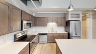 This two-story loft-style space combines industrial design elements with contemporary finishes to create a functional and visually striking interior. The main level is anchored by an open-concept layout where the kitchen and living area flow seamlessly together. The kitchen is well-appointed with dark walnut-toned shaker cabinets, white quartz countertops, a decorative tile backsplash, stainless steel appliances, and a central island with pendant lighting overhead. Exposed structural elements on the ceiling reinforce the industrial character of the space.

A standout feature of the main level is the dramatic floor-to-ceiling windows along one wall, which flood the space with natural light and offer views of the surrounding urban streetscape. A sleek open-riser staircase with metal railings and cable detailing leads to the upper level, which houses the private living quarters. The upper floor includes a spacious walk-in closet outfitted with wire shelving systems and carpeted flooring, along with two bathrooms. One bathroom features a glass-enclosed walk-in shower, a dark-cabinet vanity, and wood-look flooring. The second bathroom offers a full tub-shower combination with a matching vanity. Light wood-look flooring is consistent throughout the main living areas, unifying the overall aesthetic.