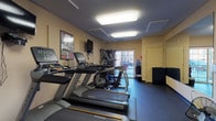 This fitness center is a dedicated, single-level exercise facility designed to support a comprehensive workout routine. The space is outfitted with a suite of Matrix-brand cardio equipment, including multiple treadmills and elliptical cross-trainers, each equipped with individual digital display consoles for personalized training. A multi-station cable and strength machine anchors one end of the room, offering a versatile range of resistance exercises. A full dumbbell rack lines one wall, providing a complete selection of free weights for strength training. A large wall-mounted mirror spans a significant portion of one wall, visually expanding the space and allowing users to monitor their form during workouts. Wall-mounted flat-screen televisions are positioned for easy viewing from the cardio equipment. The flooring consists of durable, dark rubber gym matting throughout, providing safety and cushioning. Flush-mounted fluorescent ceiling panels deliver bright, even illumination across the entire space. Large windows and glass doors along one wall introduce natural light and offer views to the outdoors. Additional amenities include a water cooler, a wall clock, exercise reference charts, and a coat hook area near the entrance. Adjacent changing rooms or restroom facilities are accessible directly from the gym floor.