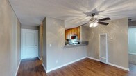 This single-floor space features an open and functional layout that seamlessly connects the living area to the kitchen. The living area is finished with warm hardwood-style flooring that extends throughout, complemented by neutral gray walls and crisp white baseboards that create a clean, cohesive aesthetic. A ceiling fan with a light fixture is centrally positioned overhead, providing both airflow and ambient lighting. The textured popcorn ceiling adds a classic touch to the space.

The kitchen is accessible via a half-wall pass-through with a dark granite-style countertop, offering a casual bar or serving area that maintains visual openness between the two spaces. Inside the kitchen, warm-toned wood cabinetry provides ample storage, while a tiled backsplash adds a decorative element. A built-in range and overhead microwave or vent hood are also present. A wall-mounted HVAC vent is conveniently located in the living area. Near the entrance, a white door with standard hardware leads into a hallway, providing a clear and practical flow throughout the single-level layout.