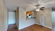 This single-floor space features an open and functional layout that seamlessly connects the living area to the kitchen. The living area is finished with warm hardwood-style flooring that extends throughout, complemented by neutral gray walls and crisp white baseboards that create a clean, cohesive aesthetic. A ceiling fan with a light fixture is centrally positioned overhead, providing both airflow and ambient lighting. The textured popcorn ceiling adds a classic touch to the space.

The kitchen is accessible via a half-wall pass-through with a dark granite-style countertop, offering a casual bar or serving area that maintains visual openness between the two spaces. Inside the kitchen, warm-toned wood cabinetry provides ample storage, while a tiled backsplash adds a decorative element. A built-in range and overhead microwave or vent hood are also present. A wall-mounted HVAC vent is conveniently located in the living area. Near the entrance, a white door with standard hardware leads into a hallway, providing a clear and practical flow throughout the single-level layout.