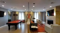 Located at 750 Summer St in Stamford, Connecticut, this single-floor amenity space is designed to serve as a dynamic and social gathering destination. The open-plan layout is anchored by a full-size pool table with a striking red felt surface, making it an immediate focal point upon entry. Opposite the billiards area, a generously sized lounge zone features a large sectional sofa in charcoal gray, accented by vibrant orange throw pillows and a bold, color-blocked orange area rug that energizes the space. Sleek pendant lighting with dark industrial-style fixtures hangs from the ceiling, complementing recessed lighting that ensures the entire area is well-illuminated. Structural columns punctuate the floor plan while adding an architectural character to the room. Floor-to-ceiling curtains frame large windows that allow natural light to filter throughout the space, creating an open and airy atmosphere. Additional seating nooks with armchairs and accent tables provide quieter spots for smaller gatherings. A decorative vintage red telephone booth serves as a playful and eye-catching design element along the back wall. The neutral gray and white color palette, punctuated by bold orange accents, gives the space a modern, curated aesthetic that is both functional and visually engaging.