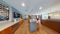 This well-appointed single-floor communal space showcases a thoughtfully designed open-plan layout that seamlessly connects multiple functional zones. The kitchen area serves as a central hub, featuring warm wood cabinetry along an extended counter wall, a large central island with bar seating, and pendant lighting overhead. Decorative laser-cut metal panels flank a wall-mounted television, adding an artistic focal point above a tiled backsplash. Stainless steel appliances and light quartz countertops provide a clean, modern contrast to the rich wood tones throughout.

The lounge area transitions naturally from the kitchen, offering a distinctive slatted wood ceiling that adds architectural depth and warmth. This section features a large flat-screen television, casual seating including rounded chairs and woven ottomans, and large windows that invite generous natural light and outdoor views. An adjacent dining nook includes a geometric shelving unit, a round table with mixed seating, and draping curtain panels that soften the space.

The main lounge area is anchored by plush swivel chairs, patterned banquette seating, and curated artwork. Warm hardwood flooring flows consistently throughout, unifying the space. The overall ambiance is contemporary and welcoming, balancing functional design with stylish, eclectic decor.