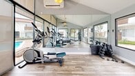 Located at 201 Los Arbolitos Blvd in Oceanside, California, this fitness center offers a bright and functional exercise space designed to support a well-rounded workout routine. The room features light wood-style flooring throughout and is bathed in natural light, thanks to expansive floor-to-ceiling glass windows and sliding doors that line the walls and open to views of the outdoor pool area and surrounding community.

The vaulted ceiling enhances the sense of openness, and ceiling fans provide comfortable airflow during workouts. A wall of mirrors visually expands the space and aids in form correction during exercise. Climate control is provided by a mounted mini-split air conditioning unit, ensuring year-round comfort.

The fitness center is well-stocked with a variety of equipment, including treadmills, an elliptical trainer, a stationary bike, and a multi-station cable machine by Hoist. A dedicated free weights area includes a dumbbell rack and a weight bench. A medicine ball storage rack rounds out the strength training options.

The space also features a large wall-mounted television, a paper towel dispenser, and a waste bin, adding convenience to the overall experience. A private entrance door leads directly to the outdoor amenity area, seamlessly connecting the fitness center to the pool and patio beyond.