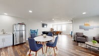 This single-floor space is designed as a versatile entertainment and social hub, thoughtfully organized into several distinct functional zones unified by rich, reclaimed-style hardwood flooring and a clean, modern aesthetic. Recessed lighting throughout ensures consistent, well-distributed illumination across the open layout.

The main gathering area features a round white dining table surrounded by mid-century modern blue upholstered chairs, anchored by a vibrant floral area rug. Adjacent to this is a relaxed lounge corner with plush gray accent chairs, a neutral area rug, and large windows fitted with white blinds that invite natural light and outdoor views.

A dedicated games zone includes a foosball table, a round poker table with leather chairs, a wall-mounted flat-screen television, and a putting green, providing diverse recreational options. French doors and multiple windows along one wall enhance the sense of openness throughout this area.

The kitchenette is neatly appointed with white shaker cabinetry, light quartz countertops, a stainless steel undermount sink with a brushed nickel faucet, and a glass-ceramic cooktop, alongside a full-size stainless steel refrigerator.

An accessible restroom features grab bars, a small privacy window, and the same hardwood-style flooring that flows throughout the rest of the space.