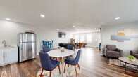 This single-floor space is designed as a versatile entertainment and social hub, thoughtfully organized into several distinct functional zones unified by rich, reclaimed-style hardwood flooring and a clean, modern aesthetic. Recessed lighting throughout ensures consistent, well-distributed illumination across the open layout.

The main gathering area features a round white dining table surrounded by mid-century modern blue upholstered chairs, anchored by a vibrant floral area rug. Adjacent to this is a relaxed lounge corner with plush gray accent chairs, a neutral area rug, and large windows fitted with white blinds that invite natural light and outdoor views.

A dedicated games zone includes a foosball table, a round poker table with leather chairs, a wall-mounted flat-screen television, and a putting green, providing diverse recreational options. French doors and multiple windows along one wall enhance the sense of openness throughout this area.

The kitchenette is neatly appointed with white shaker cabinetry, light quartz countertops, a stainless steel undermount sink with a brushed nickel faucet, and a glass-ceramic cooktop, alongside a full-size stainless steel refrigerator.

An accessible restroom features grab bars, a small privacy window, and the same hardwood-style flooring that flows throughout the rest of the space.
