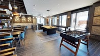 This single-floor game and social lounge is designed to serve as a dynamic communal gathering space. The open-plan layout accommodates a variety of recreational activities, anchored by a sleek black-framed billiards table with a gold felt surface, positioned at the center of the room beneath a striking horizontal linear pendant light fixture. A foosball table nearby adds to the entertainment offerings. The flooring transitions between dark wood-look tile and lighter hardwood-style planks, subtly delineating different activity zones throughout the space. A bar and dining area toward the back of the room features light cabinetry, bar-height seating, and teal accent chairs that introduce a pop of color against the otherwise neutral palette of dark grays, whites, and warm wood tones. Large floor-to-ceiling windows along the perimeter flood the interior with abundant natural light while offering views of the surrounding exterior. Wall sconces and recessed lighting complement the pendant fixtures overhead, ensuring the space is well-lit throughout. The overall ambiance is polished and contemporary, blending functionality with an upscale aesthetic suited for social gatherings and leisure.