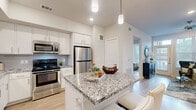 This single-floor residence showcases a thoughtfully designed open-concept layout with a neutral, contemporary aesthetic carried consistently throughout. The kitchen serves as a central highlight, featuring white shaker cabinetry, grey-veined granite countertops, a subway tile backsplash, and a full suite of stainless steel appliances including a gas range, microwave, and refrigerator. A substantial kitchen island with seating provides additional workspace and casual dining options, illuminated by sleek pendant lighting overhead.

The living area flows naturally from the kitchen and dining space, with light hardwood flooring underfoot and large windows allowing generous natural light to fill the room. A ceiling fan adds both function and character to the space.

The bedroom is calm and well-proportioned, with large windows, continued hardwood flooring, and ample room for full furniture arrangements. The connected bathroom features granite vanity countertops, white cabinetry, tile flooring, and direct access to a walk-in closet equipped with built-in shelving and hanging rods.

Throughout the residence, consistent finishes — including neutral wall tones, light hardwood flooring, and granite surfaces — create a cohesive, polished environment that balances modern design with everyday livability.