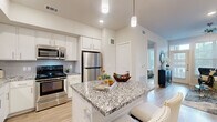 This single-floor residence showcases a thoughtfully designed open-concept layout with a neutral, contemporary aesthetic carried consistently throughout. The kitchen serves as a central highlight, featuring white shaker cabinetry, grey-veined granite countertops, a subway tile backsplash, and a full suite of stainless steel appliances including a gas range, microwave, and refrigerator. A substantial kitchen island with seating provides additional workspace and casual dining options, illuminated by sleek pendant lighting overhead.

The living area flows naturally from the kitchen and dining space, with light hardwood flooring underfoot and large windows allowing generous natural light to fill the room. A ceiling fan adds both function and character to the space.

The bedroom is calm and well-proportioned, with large windows, continued hardwood flooring, and ample room for full furniture arrangements. The connected bathroom features granite vanity countertops, white cabinetry, tile flooring, and direct access to a walk-in closet equipped with built-in shelving and hanging rods.

Throughout the residence, consistent finishes — including neutral wall tones, light hardwood flooring, and granite surfaces — create a cohesive, polished environment that balances modern design with everyday livability.