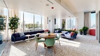 Located in Chicago's vibrant West Loop neighborhood, this contemporary office suite at 123 N Desplaines St offers a thoughtfully designed single-floor workspace that balances productivity with comfort. The layout comprises three private offices enclosed by sleek glass partitions, providing both visual openness and acoustic privacy, alongside three additional versatile spaces suited for collaboration or casual meetings.

The main lounge area is a standout feature, anchored by generous sectional seating in a calming blue tone, complemented by a mix of coral and mint accent chairs arranged around round tables — ideal for informal gatherings. Floor-to-ceiling windows wrap the perimeter, flooding the space with abundant natural light and framing striking panoramic views of the Chicago skyline and surrounding high-rises. Roller shades allow for flexible light control throughout the day.

Lush indoor plants are strategically placed throughout, adding a refreshing biophilic element to the clean, white-walled interior. The patterned carpet tile flooring ties the design together with subtle texture. Private offices are fitted with light wood furnishings and glass walls, maintaining an airy, transparent aesthetic. An adjacent outdoor terrace, visible through the windows, further enhances the sense of openness and connection to the city beyond.