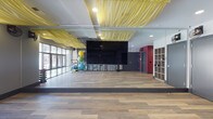 Located at 12 W Gay St in the heart of downtown Columbus, Ohio, this single-floor fitness studio offers a thoughtfully designed space built for group exercise and individual training alike. The main studio floor is expansive and open, finished with warm wood-tone flooring that provides both durability and an inviting aesthetic. A standout design element is the dramatic yellow fabric draping suspended from the ceiling, adding a bold, energetic character to the space and setting it apart from a typical gym environment. Floor-to-ceiling glass walls along one side of the studio flood the interior with natural light while maintaining a sense of openness and connectivity to the surrounding environment. A large wall-mounted display screen serves as a central focal point for instructor-led sessions or multimedia programming. Full-length mirrors along the walls enhance the sense of space and provide essential visual feedback for participants during workouts. Built-in shelving units offer convenient storage for personal items and equipment. The studio is further equipped with an assortment of fitness accessories including stability balls and other training tools, neatly stored along the perimeter. The overall ambiance is modern, vibrant, and functional, designed to energize and motivate those who use the space.
