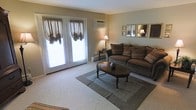 This single-floor space offers a welcoming and traditionally styled interior designed for comfortable everyday living. The living room serves as the heart of the home, featuring plush, earth-toned upholstered seating arranged around a classic dark wood oval coffee table, anchored by a layered area rug that adds warmth and definition to the space. Soft, neutral wall tones create a calm and inviting ambiance throughout. A pair of French doors dressed with draped balloon-style curtains floods the room with natural light while offering a connection to the outdoors. A wall-mounted air conditioning unit ensures year-round comfort, complemented by baseboard heating along the perimeter. Decorative framed mirrors and artwork adorn the walls, adding a refined, homey character to the space. A traditional table lamp provides soft, ambient lighting for evening hours. The carpeted flooring adds a layer of warmth underfoot, tying the room's classic aesthetic together cohesively. Overall, this space presents a tidy, well-maintained interior with a timeless and functional design sensibility.