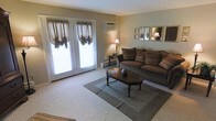 This single-floor space offers a welcoming and traditionally styled interior designed for comfortable everyday living. The living room serves as the heart of the home, featuring plush, earth-toned upholstered seating arranged around a classic dark wood oval coffee table, anchored by a layered area rug that adds warmth and definition to the space. Soft, neutral wall tones create a calm and inviting ambiance throughout. A pair of French doors dressed with draped balloon-style curtains floods the room with natural light while offering a connection to the outdoors. A wall-mounted air conditioning unit ensures year-round comfort, complemented by baseboard heating along the perimeter. Decorative framed mirrors and artwork adorn the walls, adding a refined, homey character to the space. A traditional table lamp provides soft, ambient lighting for evening hours. The carpeted flooring adds a layer of warmth underfoot, tying the room's classic aesthetic together cohesively. Overall, this space presents a tidy, well-maintained interior with a timeless and functional design sensibility.