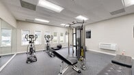 Located at 8805 Dunwoody Pl in Sandy Springs, Georgia, this fitness center offers a comprehensive, single-floor exercise space designed to support a full range of workout routines. The room features durable rubber flooring throughout, providing both comfort and safety during exercise. A drop-panel ceiling with recessed fluorescent lighting ensures bright, even illumination across the entire space.

Natural light filters in through multiple windows along one wall, offering views of the surrounding greenery and creating an open, inviting atmosphere. A full-length mirror along one wall further enhances the sense of space while allowing users to monitor their form during workouts.

The facility is well-stocked with a variety of equipment. Cardio offerings include elliptical trainers and a treadmill, while a multi-station cable machine supports a wide range of strength training exercises. An adjustable weight bench, stability balls, medicine balls, step mats, and a punching bag round out the selection, catering to diverse fitness goals. Equipment placards and informational posters are mounted on the walls for user reference.

Climate control is provided by a wall-mounted HVAC unit, ensuring year-round comfort. A keyed entry door leads to the exterior, and interior access connects to adjacent areas. The space is clean, organized, and thoughtfully laid out to maximize usability for residents.