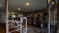 This fitness center is designed to support a wide range of workout routines in a focused, motivating environment. The space is anchored by a bold motivational mural spanning the main wall, boldly posing the question "What's Your Goal?" and filled with an array of fitness aspirations — setting an energizing tone throughout the room. Recessed lighting illuminates the dark rubberized flooring, creating a sleek, purposeful aesthetic. 

The cardio section is well-appointed with a row of Life Fitness treadmills and upright elliptical machines, each equipped with individual display screens for tracking performance. Adjacent to these, stationary bikes and other cardio equipment provide ample variety for endurance training.

The strength training area features a Smith machine, a multi-cable functional trainer, and a dedicated weight bench, complemented by a full rack of dumbbells. Along one wall, large mirrors amplify the sense of space and allow for form monitoring during workouts. 

A functional training zone is stocked with kettlebells, medicine balls, stability balls, a BOSU trainer, TRX suspension straps, and battle ropes, offering versatile options for dynamic and conditioning exercises. Natural light filters in through windows, adding a welcoming brightness to the otherwise purposefully moody interior design.