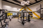 This contemporary fitness facility is designed with both functionality and visual impact in mind. The main workout floor is expansive, featuring high ceilings with exposed industrial-style elements and a dramatic open mezzanine level above, accessible via a striking black spiral staircase. The upper level is outfitted with additional cardio and training equipment, maximizing the full vertical capacity of the space.

The main floor is home to a comprehensive selection of strength training equipment, including cable machines, power racks, Smith machines, adjustable benches, and a full dumbbell rack arranged along mirrored walls that enhance the sense of openness. A dedicated "Training Zone" is clearly marked with bold yellow-and-black diagonal striped flooring and wall graphics, creating a visually distinct area for functional and free-weight training.

Geometric hexagonal pendant lights hang from the ceiling, providing focused illumination across the workout floor, while linear LED strip lighting runs throughout the space. The grey carpet tile flooring offers a durable yet comfortable surface underfoot.

A separate utility corridor leads to clean, well-lit restroom facilities with recessed overhead lighting and keycard-access doors. The facility's consistent black-and-yellow color scheme throughout the walls, flooring accents, and equipment creates a cohesive, high-energy athletic environment.