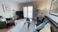This single-floor residence offers a thoughtfully designed living space that combines modern aesthetics with everyday comfort. The living room serves as the focal point of the home, showcasing light-toned hardwood-style flooring that extends throughout and contributes to a clean, contemporary feel. A large sliding glass door dominates one wall, fitted with vertical blinds that allow for adjustable natural light and provide a view to the outdoors, creating an open and airy atmosphere. The space is furnished with a sleek leather sofa accented by teal throw pillows, complemented by a round glass-top coffee table with a sculptural wooden base resting on a patterned area rug. A flat-screen television is positioned on a low media console in the corner, while a tall arc floor lamp and a curated gallery wall featuring botanical and nature-inspired artwork add character and warmth to the neutral wall tones. A lush indoor plant enhances the room's natural ambiance. The overall design palette — neutral walls, soft textiles, and carefully chosen accent pieces — creates a cohesive, inviting environment that is both functional and visually appealing, making this a comfortable and stylish single-level home.