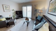 This single-floor residence offers a thoughtfully designed living space that combines modern aesthetics with everyday comfort. The living room serves as the focal point of the home, showcasing light-toned hardwood-style flooring that extends throughout and contributes to a clean, contemporary feel. A large sliding glass door dominates one wall, fitted with vertical blinds that allow for adjustable natural light and provide a view to the outdoors, creating an open and airy atmosphere. The space is furnished with a sleek leather sofa accented by teal throw pillows, complemented by a round glass-top coffee table with a sculptural wooden base resting on a patterned area rug. A flat-screen television is positioned on a low media console in the corner, while a tall arc floor lamp and a curated gallery wall featuring botanical and nature-inspired artwork add character and warmth to the neutral wall tones. A lush indoor plant enhances the room's natural ambiance. The overall design palette — neutral walls, soft textiles, and carefully chosen accent pieces — creates a cohesive, inviting environment that is both functional and visually appealing, making this a comfortable and stylish single-level home.