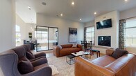 Located at 950 Brushfield Dr in Galloway, Ohio, this single-story community clubhouse showcases a contemporary, open-concept design with light hardwood-style flooring and recessed lighting throughout. The expansive lounge area serves as the heart of the space, anchored by a floor-to-ceiling white brick fireplace with a mounted flat-screen TV above. Comfortable leather sofas and upholstered wingback chairs are thoughtfully arranged to encourage both relaxation and social gathering, complemented by layered area rugs and curated wall art. Large windows dressed with patterned drapes allow abundant natural light to fill the interior, creating a bright and welcoming ambiance. Adjacent to the lounge, a dedicated dining area with bar-height seating provides a casual spot for residents to gather. The well-appointed kitchen features a striking two-tone design with crisp white upper cabinetry, dark lower cabinetry, white quartz countertops, a subway tile backsplash, stainless steel appliances, and a central island with pendant lighting overhead. A glass-walled private workspace is seamlessly integrated into the layout, offering a quiet, professional environment visible from the main lounge. The overall design balances modern sophistication with a warm, approachable character suited for community living.
