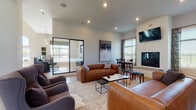 Located at 950 Brushfield Dr in Galloway, Ohio, this single-story community clubhouse showcases a contemporary, open-concept design with light hardwood-style flooring and recessed lighting throughout. The expansive lounge area serves as the heart of the space, anchored by a floor-to-ceiling white brick fireplace with a mounted flat-screen TV above. Comfortable leather sofas and upholstered wingback chairs are thoughtfully arranged to encourage both relaxation and social gathering, complemented by layered area rugs and curated wall art. Large windows dressed with patterned drapes allow abundant natural light to fill the interior, creating a bright and welcoming ambiance. Adjacent to the lounge, a dedicated dining area with bar-height seating provides a casual spot for residents to gather. The well-appointed kitchen features a striking two-tone design with crisp white upper cabinetry, dark lower cabinetry, white quartz countertops, a subway tile backsplash, stainless steel appliances, and a central island with pendant lighting overhead. A glass-walled private workspace is seamlessly integrated into the layout, offering a quiet, professional environment visible from the main lounge. The overall design balances modern sophistication with a warm, approachable character suited for community living.
