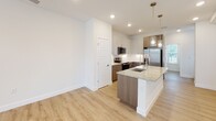 This two-story space showcases a thoughtfully designed interior with contemporary finishes throughout. The main level centers around an open kitchen equipped with a large granite-topped island featuring a double undermount sink and pendant lighting above. Cabinetry is presented in a two-tone combination of white upper cabinets and warm walnut-toned lower cabinets, paired with a clean white subway tile backsplash. Stainless steel appliances, including a French door refrigerator, electric range, and microwave, complete the kitchen. Light wood-tone luxury vinyl plank flooring runs throughout the main living areas, creating a bright, cohesive aesthetic against crisp white walls.

A half bathroom on the main level includes a granite vanity top with an undermount sink and a window providing natural light. The upper level hosts carpeted bedrooms with ceiling fans and windows overlooking greenery. A full bathroom features a double-sink granite vanity and a glass-enclosed walk-in shower with large-format stone-look tile. A dedicated laundry room offers a full-size washer and dryer alongside ample cabinetry and a granite-topped utility sink, adding exceptional functionality to the space.