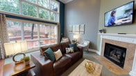 This well-appointed clubhouse offers a thoughtfully designed single-floor layout that immediately impresses with its soaring ceilings and dramatic floor-to-ceiling multi-pane window wall. The oversized windows flood the interior with abundant natural light and provide sweeping views of the surrounding mature trees and landscaped exterior, creating a bright and inviting atmosphere throughout the day.

The main lounge area serves as the heart of the clubhouse, anchored by a classic white-mantle fireplace with elegant tile surround and a wall-mounted flat-screen television above. Rich hardwood flooring at the entry transitions seamlessly into plush carpeting in the lounge, adding warmth and texture to the space. A rich brown leather sofa, coordinating accent chairs, and wood coffee tables are arranged for comfortable gathering and conversation.

Deep teal accent walls contrast beautifully with the soft gray tones throughout, lending the space a sophisticated yet welcoming character. Crown molding and crisp white baseboards add refined architectural detail. French doors provide convenient direct access to the outdoor courtyard, enhancing the indoor-outdoor connection. Decorative artwork, ambient table lamps, and curated accessories complete the polished, community-focused aesthetic of this clubhouse.