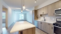 Located at 4560 Mission Gorge Pl in San Diego, CA, this single-floor residence offers a thoughtfully designed layout with modern finishes throughout. The open-concept kitchen serves as the heart of the space, showcasing light wood-grain cabinetry with matte black hardware, a white quartz countertop island with pendant lighting overhead, a subway tile backsplash, and a full suite of stainless steel appliances including a gas range, built-in microwave, dishwasher, and refrigerator. A matte black faucet at the sink adds a refined contrast to the warm-toned cabinetry. The kitchen flows seamlessly into the living area, which features durable wood-look flooring, a ceiling fan, and a large window that fills the room with natural light. The bedroom is carpeted for comfort and includes a window with blinds offering ample natural light. The full bathroom is finished with large-format tile flooring, a wood-grain vanity with white countertop and matte black fixtures, a mirrored medicine cabinet, and a tub-shower combo enclosed by a sliding glass door. A compact private outdoor area with tiled flooring completes the space, providing a quiet exterior retreat. The unit's cohesive design palette of warm wood tones, white surfaces, and black accents creates a clean, contemporary aesthetic throughout.