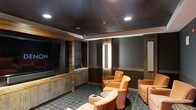 This single-level screening room is designed to deliver a premium home theater experience through a carefully curated blend of materials, lighting, and furnishings. The space is anchored by a large wall-mounted display set within a rich wood-paneled surround, complemented by built-in cabinetry below that provides both storage and a serving surface — ideal for entertaining. The dark, coffered ceiling with recessed lighting creates a dramatic overhead canopy that enhances the room's immersive atmosphere while minimizing glare on the screen. Vertical illuminated accent panels along the side walls add a warm, ambient glow reminiscent of a high-end commercial cinema. The seating consists of generously sized leather recliners in a warm cognac tone, arranged in a forward-facing configuration with integrated cup holders for convenience. The flooring features a patterned carpet in cool gray tones, which absorbs sound and contributes to the room's acoustic quality. A projection screen is also visible along the right wall, offering versatility for larger-format viewing. Overall, the space balances refined aesthetics with functional design.