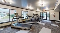 Located at 1735 Ashley Hall Rd in Charleston, SC, this fitness center is a single-floor facility designed to support a comprehensive workout experience. The space features a patterned carpet floor with designated white workout zones that help organize equipment areas and provide dedicated space for floor exercises.

The cardio section is equipped with multiple treadmills and elliptical machines, positioned along windowed walls that allow natural light to fill the room. Large mirrors on one wall enhance the sense of space while allowing users to monitor their form. A glass door at one end of the room opens to an outdoor area, further contributing to the bright and open atmosphere.

The strength and functional training area includes an adjustable workout bench, a rack of dumbbells in varying weights, and resistance bands mounted on the wall for versatile training options. A black foam pad is also available for stretching and floor work.

Motivational framed wall art featuring athletic silhouettes and bold typography—reading "Harder, Faster, Stronger"—adds an energizing visual element to the space. Track lighting and ceiling-mounted fixtures provide consistent illumination throughout. The overall design is clean, functional, and purposefully arranged to accommodate a variety of fitness routines.