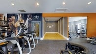 VIE Fit at Murfreesboro is a well-appointed fitness center designed to inspire and motivate. The space is defined by a bold, contemporary aesthetic, featuring a striking color palette of deep gray and vibrant orange accents that create an energetic atmosphere throughout. A large, eye-catching branded mural anchors one wall, reinforcing the identity of the facility and setting an aspirational tone for members.

The fitness center is thoughtfully divided into distinct functional zones. The cardio area is equipped with modern machines, including elliptical trainers, positioned on durable, cushioned flooring ideal for high-traffic use. Adjacent to this, a dedicated open training corridor — finished with smooth hardwood-style flooring — provides ample space for functional fitness, stretching, or group movement exercises. This area is clearly marked with the "VIE FIT" branding, making it a central visual focal point of the space.

Further into the facility, a strength training section houses cable machines and resistance equipment, catering to a full range of workout needs. Recessed lighting throughout ensures consistent, bright illumination across all areas. A separate glass-door room offers additional private or specialized training space. The overall design balances practicality with a polished, gym-quality environment suited for a diverse range of fitness routines.