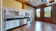 This loft-style space showcases a compelling blend of industrial heritage and modern updates. The open main living area immediately draws the eye with its soaring ceilings, original heavy timber columns and beams, exposed brick accent walls, and large windows that invite abundant natural light and outdoor views. Polished hardwood-style flooring runs throughout, complementing the warm, industrial aesthetic created by the exposed spiral ductwork and track lighting overhead.

The kitchen is well-appointed with white shaker-style cabinetry, dark stone countertops, and a full suite of stainless steel appliances, including a refrigerator, electric range, over-the-range microwave, and dishwasher. An adjacent brick wall and timber beam add architectural character to the cooking and dining area.

The bathroom is finished with matching white cabinetry, a dark stone vanity top, a large mirror with overhead lighting, and hardwood-style flooring, maintaining a cohesive design throughout. A dedicated laundry room provides practical utility with an in-unit washer and storage shelving, along with a water heater and HVAC equipment housed in the same space.

Overall, this single-floor space offers a distinctive urban loft ambiance, balancing the raw character of its historic building origins with thoughtful modern finishes.