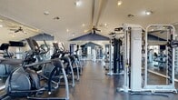 This fitness center offers a spacious, single-floor layout thoughtfully designed to accommodate a comprehensive workout experience. The room is defined by its striking vaulted ceiling with a bold navy blue accent that follows the roofline's peak, adding architectural character and a sense of height to the space. Recessed lighting and ceiling fans are positioned throughout, ensuring the area remains well-lit and well-ventilated during use.

Large floor-to-ceiling mirrors line the walls, visually expanding the space and allowing users to monitor their form during exercise. The flooring consists of durable dark rubber tiles suited for high-traffic fitness use. Natural light filters in through multiple windows, including a notable bay window in the free weights area, providing views of the surrounding exterior.

The cardio zone is well-stocked with treadmills, elliptical machines, and stationary bikes, while the strength training area includes a multi-station cable machine, a Smith machine or power rack, adjustable benches, and a full dumbbell rack with a range of weights. Wall-mounted flat-screen televisions are positioned for easy viewing from the equipment. The overall ambiance is clean, functional, and motivating, making this fitness center a practical and well-appointed facility for daily training.