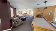 This inviting single-floor recreational space is designed for leisure and social activity, offering a well-appointed game room and lounge area. The open layout is anchored by multiple solid wood pool tables with classic leather pockets, each illuminated by stylish pendant light fixtures that hang above. The room is finished with dark patterned carpeting throughout, complementing the warm yellow and cream-toned walls accented by rich dark burgundy structural columns that add architectural character.

Natural light flows generously into the space through large windows and French doors that line one side of the room, creating a bright and welcoming atmosphere. A comfortable seating arrangement featuring patterned accent chairs and a tufted leather ottoman provides a relaxed lounge area ideal for conversation or spectating. Decorative wall art adds a contemporary touch to the space.

Additional amenities include a wall-mounted cue rack, a bar-height shelf with seating, and access to an adjacent kitchenette or bar area visible through a connecting passageway. Recessed and pendant lighting throughout ensures the space is well-lit at all times. The combination of recreational features, lounge seating, and ample natural light makes this a versatile and comfortable communal gathering space.