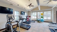 This single-floor community clubhouse offers a thoughtfully designed and welcoming environment suited for both relaxation and active living. The main entrance opens into a bright, airy lobby with soaring ceilings, warm hardwood-trimmed double doors, and elegant chandelier lighting. Natural light pours in through large windows, creating an inviting atmosphere throughout the space.

The central lounge area is furnished with a tufted cream sofa, accent chairs, and a dining table, offering flexible seating arrangements for socializing or informal gatherings. Black-framed French doors with glass panels divide the lounge into distinct zones while maintaining an open, connected feel. Soft neutral tones, textured area rugs, and carefully selected décor contribute to a refined yet comfortable ambiance.

A dedicated sitting area anchored by a striking floor-to-ceiling stone fireplace with a rustic wood mantel serves as a cozy focal point. French doors adjacent to the fireplace open to an outdoor area, allowing seamless indoor-outdoor flow and filling the room with natural light.

The on-site fitness center is well-equipped with treadmills, an elliptical, a stationary bike, and an extensive selection of free weights, resistance equipment, and exercise balls. Vaulted ceilings with a ceiling fan, multiple windows, and a wall-mounted television enhance the workout environment, making it both functional and comfortable.