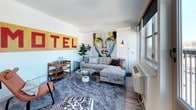 This contemporary single-floor unit showcases a thoughtfully curated design that blends modern finishes with eclectic, art-forward décor throughout. The living area is a standout space, anchored by a large sectional sofa, a glass coffee table, and an eye-catching large-scale portrait painting. A bold "MOTEL" sign mounted above a mid-century bar cart adds a playful, urban personality to the room. Generous windows with wood-trimmed roller shades flood the space with natural light and offer sweeping city rooftop views, enhancing the open, airy ambiance. The kitchen features a sleek two-tone design with dark lower cabinets and white upper cabinetry, complemented by stainless steel appliances including a range, over-the-range microwave, and dishwasher, along with a modern undermount sink. The bedroom is bright and welcoming, with a large window framing expansive city views, light wood-style flooring, and thoughtfully placed artwork that adds character to the clean white walls. The bathroom offers a well-appointed vanity with an undermount sink, quartz-style countertop, a large wood-framed mirror, and modern cabinetry, all accented by framed art pieces that carry through the space's distinctive aesthetic.