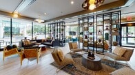 Located at 1000 Lake St in Oak Park, Illinois, this thoughtfully designed lobby level offers a welcoming and visually dynamic environment that blends contemporary architecture with eclectic interior design. The entry foyer greets visitors with light wood herringbone-patterned flooring, ornate ceiling detailing, and floor-to-ceiling glass walls that flood the space with natural light and provide clear sightlines to the surrounding streetscape.

The main lounge area is generously proportioned, furnished with a curated mix of seating including curved velvet sofas, patterned accent chairs, and layered area rugs that create distinct gathering zones. Open black metal shelving units display decorative objects, adding an artful, boutique-like character throughout. A dedicated lounge nook features a recessed linear fireplace set against a richly textured wood-paneled wall, complemented by geometric-patterned drapery and accent lighting.

A glass-partitioned area with a wood-slat ceiling houses the leasing or management office, offering a professional yet design-forward workspace visible from the main corridor. A living green wall installation punctuates one section, adding a natural element amid the layered wood tones and dark accents. Tufted banquette seating and pendant lighting define a cozy social corner, rounding out the variety of spaces available within this well-appointed lobby.