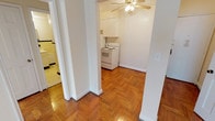 This single-floor residence is defined by its consistent use of warm herringbone parquet flooring, which extends throughout the living areas, bedroom, and hallway, lending a classic, cohesive character to the space. The layout, as seen in the overhead model, comprises a spacious main living area, a separate bedroom, a galley-style kitchen, and a full bathroom.

The living and bedroom areas are generously proportioned, each featuring ceiling fans with integrated lighting and large windows that allow natural light to fill the rooms. Radiator heating units are positioned beneath the windows in both spaces. The walls are finished in a clean, neutral white, complementing the warm tones of the parquet floors.

The galley kitchen is efficiently arranged with white cabinetry, a stainless steel sink, laminate countertops, and white appliances including a coil-burner range and a full-size refrigerator. The parquet flooring continues into this area, maintaining visual continuity.

The bathroom features a classic design with cream and black square wall tiles, a basket-weave tile floor, a full soaking tub with an overhead shower, and a towel bar — all contributing to a timeless, vintage aesthetic throughout the unit.