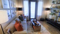 Located in Flower Mound, Texas, this single-floor space showcases a thoughtfully designed living area that balances comfort with contemporary style. The living room serves as the centerpiece of the space, anchored by a plush, light-toned sofa dressed with an array of accent pillows in warm rust and cool gray tones, lending a sophisticated yet welcoming atmosphere.

A woven rectangular coffee table sits atop a softly patterned area rug, adding texture and visual interest to the room. Hardwood-style flooring extends throughout, contributing warmth and cohesion to the overall aesthetic. A full-length glass door flanked by sheer white curtains draws in natural light while offering access to an outdoor area, enhancing the sense of openness within the space.

Ambient lighting is thoughtfully incorporated through sleek floor and table lamps positioned on either side of the seating area, creating a warm, layered glow. Framed artwork adorns the walls, adding a curated, gallery-like touch. The overall ambiance is cozy and refined, reflecting a modern design sensibility with carefully selected furnishings and décor that make the space feel both livable and polished.