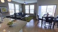 This single-floor space showcases a well-appointed living room designed with a cohesive, contemporary aesthetic. The room is anchored by a plush, light gray sofa adorned with decorative pillows featuring a subtle leaf motif, complementing the nature-inspired drapery panels that frame the windows. A glass-top coffee table with a sleek black metal frame serves as the room's focal point, resting atop a textured area rug that adds warmth and definition to the space. A cream accent chair positioned near the window provides an inviting reading nook bathed in natural light filtered through horizontal blinds. The walls are finished in a warm taupe tone, creating a calm and inviting atmosphere. Soft ambient lighting is provided by a classic white table lamp, while a large framed artwork on the wall adds a refined artistic touch. The overall design balances functionality and style, with carefully selected décor pieces contributing to a polished, livable environment. Natural light from multiple windows enhances the sense of openness throughout the room.