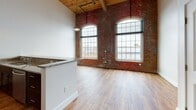 Located at 89 Quincy Ave in Pawtucket, Rhode Island, this two-floor loft-style residence is a thoughtfully converted mill building that masterfully blends industrial heritage with modern finishes. The main living level immediately impresses with its soaring wood-beamed ceilings, exposed brick walls, and dramatic arched windows that frame city views while allowing abundant natural light to pour in. Visible industrial ductwork runs overhead, reinforcing the building's authentic mill character. Warm hardwood flooring extends throughout both levels, lending continuity and warmth to the space.

The open kitchen is outfitted with rich, dark cabinetry, granite countertops, stainless steel appliances, and a peninsula that defines the boundary between cooking and living areas. The upper floor offers a generous open area with multiple closet doors providing ample storage, and the same warm hardwood flooring continues seamlessly underfoot beneath a wood-paneled ceiling.

The bathroom showcases a refined aesthetic, featuring a patterned mosaic tile surround in neutral tones encasing a full soaking tub, wood-plank-style tile flooring, and a clean white vanity beneath a wood-paneled ceiling. A central staircase with wrought iron railings connects the two levels, completing this distinctive live-work-inspired space.