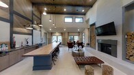 This single-floor space exudes a refined, rustic-modern aesthetic, blending natural materials with contemporary design elements to create a welcoming and stylish gathering environment. The most striking architectural feature is the soaring ceiling, accented by a rich wood-paneled section and bold black steel beams, giving the space a dramatic sense of height and openness. A row of clerestory windows allows abundant natural light to filter in from above, complementing the warm glow of the pendant light fixtures that hang gracefully over the expansive live-edge wood bar. The bar, supported by an industrial-style navy blue base and lined with black swivel stools, serves as a central focal point and functional gathering hub. To one side, a comfortable lounge area is furnished with rich leather seating, including tufted ottomans and club chairs arranged around a sculptural round coffee table. A floor-to-ceiling modern fireplace anchors the lounge, flanked by a curated stack of natural log storage that adds rustic texture and charm. A large flat-screen television is mounted above the fireplace mantle. Beyond the lounge, additional seating areas near large windows offer inviting spaces for conversation or relaxation, bathed in natural light with views to the outdoors. Light hardwood-style flooring ties the entire space together with a clean, cohesive finish.