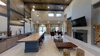 This single-floor space exudes a refined, rustic-modern aesthetic, blending natural materials with contemporary design elements to create a welcoming and stylish gathering environment. The most striking architectural feature is the soaring ceiling, accented by a rich wood-paneled section and bold black steel beams, giving the space a dramatic sense of height and openness. A row of clerestory windows allows abundant natural light to filter in from above, complementing the warm glow of the pendant light fixtures that hang gracefully over the expansive live-edge wood bar. The bar, supported by an industrial-style navy blue base and lined with black swivel stools, serves as a central focal point and functional gathering hub. To one side, a comfortable lounge area is furnished with rich leather seating, including tufted ottomans and club chairs arranged around a sculptural round coffee table. A floor-to-ceiling modern fireplace anchors the lounge, flanked by a curated stack of natural log storage that adds rustic texture and charm. A large flat-screen television is mounted above the fireplace mantle. Beyond the lounge, additional seating areas near large windows offer inviting spaces for conversation or relaxation, bathed in natural light with views to the outdoors. Light hardwood-style flooring ties the entire space together with a clean, cohesive finish.