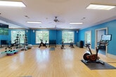 This single-story fitness center is thoughtfully organized across several interconnected areas, each designed to support a different aspect of physical training. The space is unified by a bold blue accent color along the walls, white crown molding, and flush-mounted lighting panels that provide consistent illumination throughout.

The cardio zone features multiple treadmills and stair-climber machines arranged in rows on durable rubberized flooring. Ceiling fans help maintain airflow, while a wall-mounted flat-screen TV offers entertainment during workouts. Arched windows allow natural light and outdoor views into this area.

A dedicated group exercise studio offers a spacious, open layout with light hardwood flooring, floor-to-ceiling mirrors, and a ceiling-mounted display screen. This room accommodates spin bikes, stability balls, medicine balls on a rack, step platforms, kettlebells, and stacked exercise mats, making it well-suited for a variety of group or individual classes.

A smaller auxiliary room provides additional training options, including rowing machines and wall-to-wall mirrors. Its arched windows frame views of surrounding greenery, enhancing the bright, airy feel of the space. Together, the rooms form a comprehensive, professionally outfitted fitness facility.