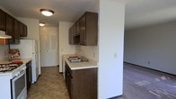 This single-floor space offers a practical and functional layout designed for comfortable everyday living. The kitchen is a standout feature, equipped with dark brown cabinetry that provides ample upper and lower storage space. A stainless steel sink is set into a light-colored countertop, offering a clean contrast to the rich tone of the cabinets. The kitchen flows openly into the adjacent living area, which is finished with wall-to-wall carpeting that adds a sense of warmth and comfort underfoot. Tile flooring in the kitchen and entryway provides a durable, easy-to-clean surface in the higher-traffic zones. The layout is thoughtfully organized, with a closet featuring louvered doors visible near the entryway, adding practical storage solutions. Flush-mount ceiling light fixtures illuminate the space throughout, keeping the interior bright and well-lit. White walls throughout the unit create a neutral, clean backdrop that allows for easy personalization. The open sightlines between the kitchen and living space contribute to an airy, connected feel, making the overall layout efficient and easy to navigate.