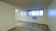 This single-floor space offers an open, functional layout characterized by clean white walls and neutral-toned carpeted flooring. The main living area is generously proportioned, featuring a bank of large windows fitted with horizontal blinds that allow ample natural light to fill the room while providing views of the surrounding trees outdoors. A window-mounted air conditioning unit is positioned along the exterior wall, complementing the baseboard heating system that runs the length of the room — providing year-round climate control options. The space connects to an adjoining area, visible through a wide open passage, which also benefits from natural light via its own window. A ceiling fan is installed in this secondary area, adding additional air circulation. The textured ceiling and straightforward architectural design contribute to a no-frills, practical aesthetic throughout. The layout offers flexibility for various furniture arrangements, and the neutral finishes provide a blank canvas that can be easily personalized. Overall, the space presents a functional and well-lit environment suitable for comfortable everyday living.