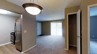 This single-floor residential space offers a practical and comfortable layout designed for everyday living. The main living area is a highlight of the space, featuring wall-to-wall plush carpeting that adds warmth underfoot and a soft, inviting ambiance throughout. A large sliding glass door fitted with vertical blinds serves as the focal point of the room, allowing generous amounts of natural light to fill the space while also providing direct access to the outdoors. The neutral, earth-toned walls in a warm taupe palette create a cohesive and understated aesthetic that complements the overall design. A decorative flush-mount ceiling light fixture anchors the room from above, providing functional overhead illumination. A dedicated closet space with an open rod system offers organized storage, keeping the living area uncluttered and efficient. The front entryway is clearly defined, featuring a white paneled door and a welcome mat, establishing a clean and practical point of entry. The textured ceiling adds a subtle architectural detail that carries throughout the space. Overall, this residence presents a straightforward, functional design with key features that support comfortable day-to-day living.